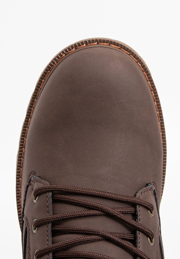 Zapato de cuero marrón con punta redondeada, costuras contrastantes y cordones marrones oscuros. Presenta una superficie texturada y un borde de suela de goma.