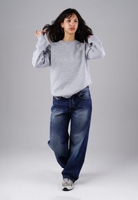Grau meliertes Sweatshirt mit Rundhalsausschnitt, lockere Passform, kombiniert mit weiten blauen Jeans. Das Model trägt silberne Creolen und sportliche graue Sneakers.