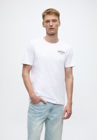 White cotton t-shirt with short sleeves featuring a black print that says "SUPERIOR CLOTHIER" on the left chest. Worn with light blue jeans.