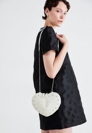Vrouw in een zwarte jurk met textuur, die een witte hartvormige tas met zilveren studs en een kettingriem over de schouder houdt, tegen een effen achtergrond.