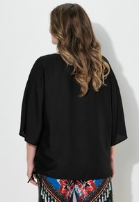 Top leggero nero con maniche ampie, caratterizzato da una vestibilità morbida e un orlo che si lega ai lati. Abbinato a pantaloni colorati e fantasia.