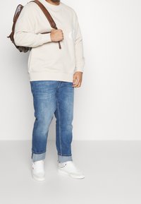 Beige Sweatshirt mit gerippten Bündchen, kombiniert mit hellblauen Jeans. Weiße Sneaker und ein brauner Leder-Rucksack runden das Outfit ab.