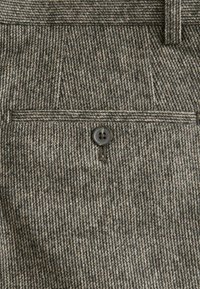 Gray and beige houndstooth fabric with a front pocket, featuring a single black button closure and textured surface.