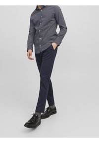 Bawełniane spodnie w kolorze granatowym i wzorzysta koszula zapinana na guziki w odcieniach niebieskiego. Czarne buty na sznurowadła. Model stoi z rękami w kieszeniach.