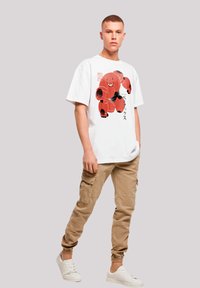 White graphic t-shirt featuring a red character design, paired with tan cargo pants and white sneakers, standing against a plain background.
