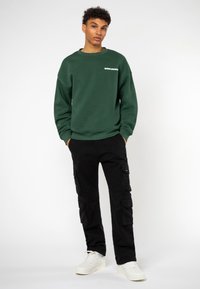 Groene crewneck sweatshirt met witte tekst, gecombineerd met zwarte cargobroek met zijzakken, en witte sneakers. Eenvoudige en relaxte pasvorm.