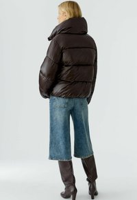 Brauner Puffy-Jacke mit hohem Kragen, gesteppter Optik und glänzender Oberfläche, kombiniert mit kurzen Jeans und kniehohen braunen Stiefeln.