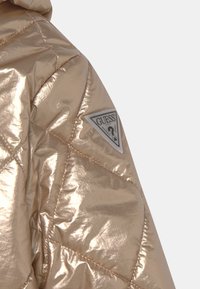 Metallic guld pufferjakke med quiltet design, struktureret overflade og et trekantet gråt logo-patch på ærmet.