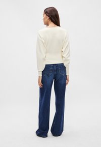 Κρεμ καλοκαιρινό πλεκτό πουλόβερ με φαρδιά μανίκια, συνδυασμένο με ψηλόμεσα, σκούρα μπλε παντελόνια με φαρδιά πόδια, διαθέτοντας πίσω τσέπες και λεπτομέρειες ραψίματος.