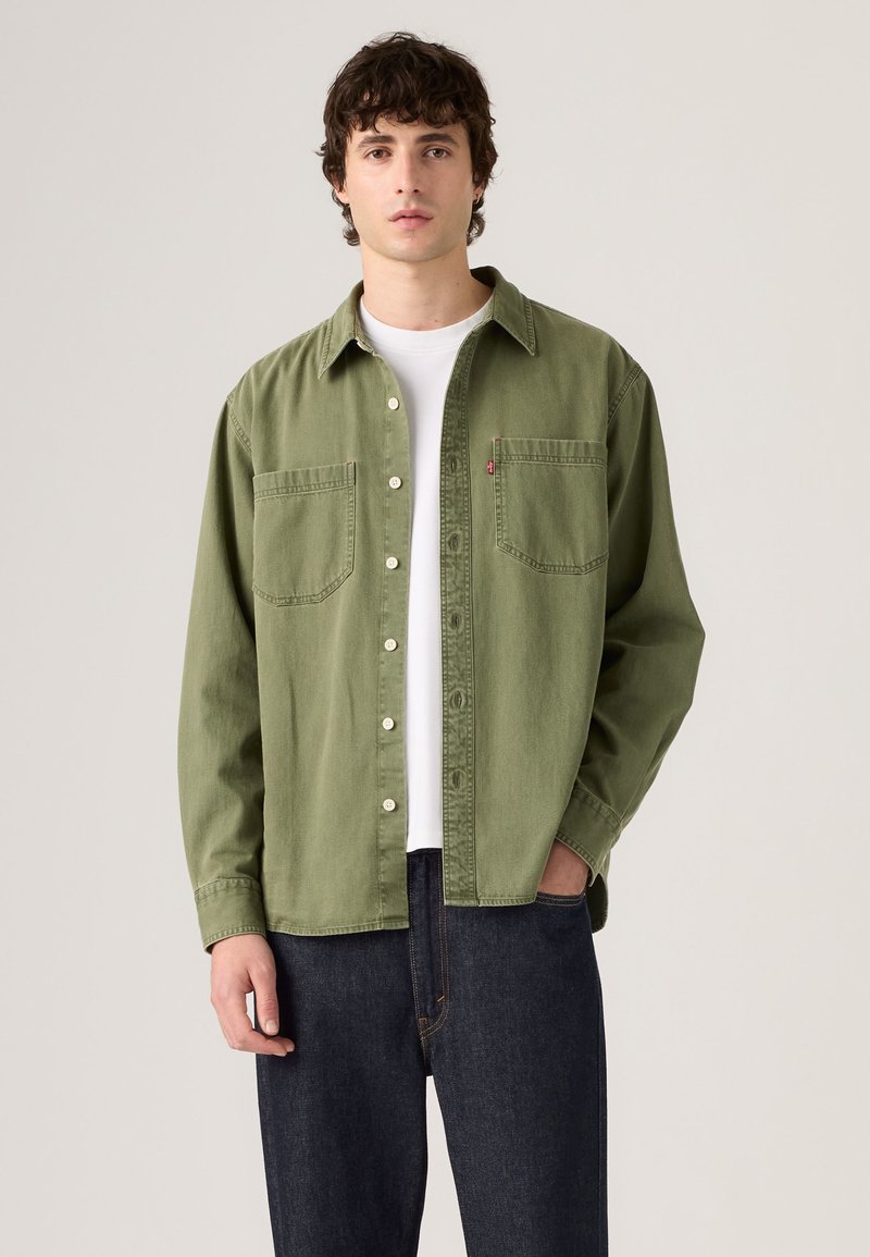 Giovane uomo con camicia verde oliva aperta sopra una t-shirt bianca e jeans blu scuro su uno sfondo chiaro e semplice.