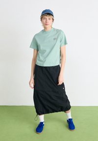 T-shirt courte vert menthe avec un logo ; jupe noire mi-longue avec ourlet à cordon de serrage ; chaussettes blanches ; baskets bleues ; fond minimaliste.