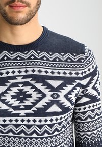 Mörkblå stickad tröja med ett vitt geometriskt mönster som innehåller diamanter och trianglar, rund halsringning och ribbade manschetter.