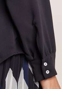 Blusa nera con maniche lunghe, tessuto arricciato al polso e due bottoni bianchi. Texture liscia, abbinata a una gonna con motivo geometrico.