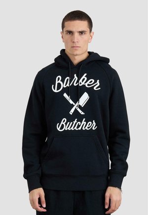 Sort hættetrøje med teksten "Barber Butcher" og grafikken med krydsede knive. Har kængurulomme og ribbede manchetter. Blød stofstruktur.