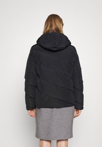 Chaqueta de abrigo negra con capucha, que presenta patrones de acolchado diagonal. Material suave y brillante con un corte holgado. Llevada sobre una falda gris.