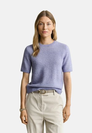 Frau mit langen blonden Haaren, die einen hellvioletten kurzärmeligen Pullover und beige Hose mit Gürtel trägt, steht vor einem schlichten Hintergrund.
