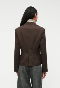 Brūns piegriezts žaketē, kas izgatavota no gludas auduma, ar pieguļošu siluetu, izgrieztiem laktiem un smalku šuvju detaļu, kombinēta ar pelēkiem platām biksēm.