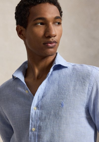 Jeune homme aux cheveux courts et bouclés portant une chemise à carreaux bleu clair à boutons, regardant vers la droite sur un fond uni.