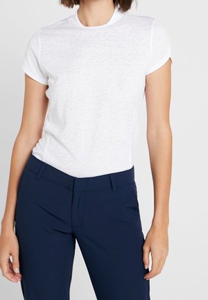 Frau trägt ein eng anliegendes weißes T-Shirt mit kurzen Ärmeln und dunkelblaue Hosen, steht mit entspannt an den Seiten hängenden Armen.