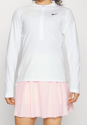 Vrouw draagt een witte Nike long-sleeve ritsvest en een lichtroze plissérok, zichtbaar van schouders tot halverwege de dijen.