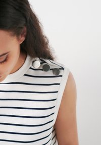 Top sin mangas blanco con rayas horizontales azul marino, que presenta cuatro botones metálicos decorativos en el hombro como detalle. Textura de tela suave.