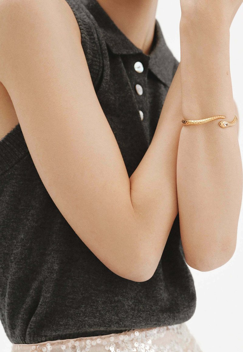 Polo sin mangas de color gris oscuro con cuello y botones, combinado con una pulsera de oro texturizada que presenta un detalle de diseño sutil.