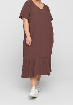 Person iført en løs brun midi-kjole med korte ærmer og hvide sneakers på en ensfarvet lys baggrund.