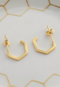 Guld hexagonala hängeörhängen med en polerad finish, med en postbakdel. Uppställda mot en tallrik med ett hexagonalt mönster.