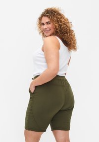 Olive zaļas šortos ar elastīgu jostu un sānu kabatām, izgatavoti no mīksta auduma. Valkāti ar baltu bez piedurknēm topu, rādīti no sānu skatpunkta.