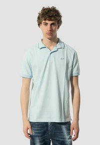 Polo shirt azzurro chiaro in cotone, con colletto classico, maniche corte e un piccolo logo sul lato sinistro del petto. Indossato con jeans blu strappati.