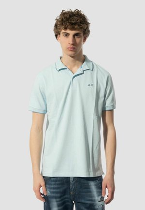 Polo shirt azzurro chiaro in cotone, con colletto classico, maniche corte e un piccolo logo sul lato sinistro del petto. Indossato con jeans blu strappati.