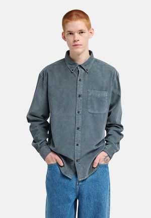Young man with short red hair wearing a gray button-up shirt and blue jeans, standing with hands in pockets against a plain background.