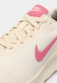 Μπεζ αθλητικό παπούτσι με υφασμένο επάνω μέρος, ροζ λογότυπο Nike και κορδόνια που ταιριάζουν. Διαθέτει στρογγυλεμένη μύτη και μαλακή σόλα.