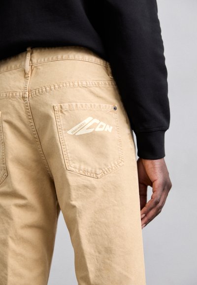 Pantalons en coton beige avec deux poches à l'arrière, ornés d'un logo blanc et de coutures. Le porteur porte une chemise noire à manches longues.