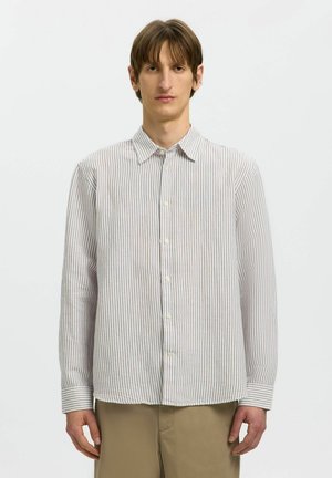 Homme portant une chemise blanche à manches longues boutonnée avec de fines rayures verticales grises et un pantalon beige, debout devant un fond blanc uni.