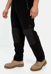 Sorte fleece-joggingbukser med kontrasterende sorte knæpaneler og en lynlåslomme. Sammen med lysebrune snørebåndsstøvler.