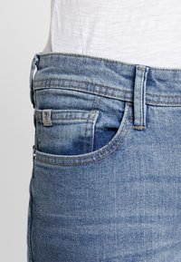 Detailní záběr osoby nosící světlé modré džínové kalhoty s přední kapsou a malou cedulkou, doplněné o bílou košili zastrčenou dovnitř.