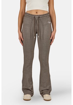 Persona con pantalones marrones de punto acanalado de cintura alta con cordón, combinados con zapatos beige y una camiseta blanca corta, de pie sobre fondo blanco.