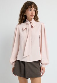 Blusa rosa pallido con un fiocco al collo, maniche lunghe a sbuffo e una texture morbida. Abbinata a pantaloni corti a vita alta color marrone scuro.