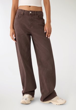 Donna che indossa pantaloni marroni a vita alta e gamba larga, top corto bianco e sneakers beige, in piedi su sfondo bianco.