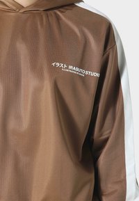 Bruine hoodie met witte zijpanelen, een gladde textuur en grafische tekst in het wit met de tekst "IRASUTO STUDIO" en Japanse karakters.