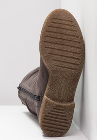 Botte en daim marron avec une semelle en caoutchouc texturée et une fermeture éclair latérale, vue de la semelle reposant contre une surface blanche.