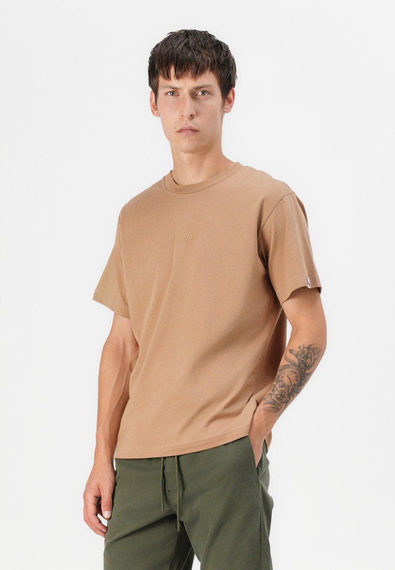 T-shirt in cotone marrone con maniche corte, scollo rotondo e logo del brand ricamato sul petto. Indossato con pantaloni verde con coulisse.