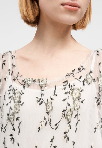 Tissu léger et transparent avec une broderie florale complexe en vert et noir, présentant un décolleté carré et des bords délicatement froncés.