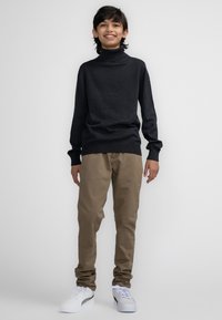 Maglione dolcevita grigio scuro, pantaloni beige e sneakers bianche. L'outfit presenta una vestibilità rilassata e polsini a coste sul maglione.