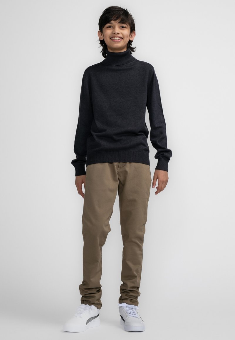 Maglione dolcevita grigio scuro, pantaloni beige e sneakers bianche. L'outfit presenta una vestibilità rilassata e polsini a coste sul maglione.