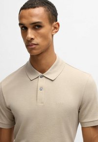 Beige polo skjorta i texturerad tyg, med krage, knäppning med tre knappar och liten broderad logotyp på bröstet.