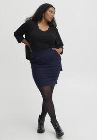 Blazer noir sur un top noir avec des bordures en dentelle, associé à une jupe crayon bleu marine et des bottines noires, présentant une texture lisse et une coupe ajustée.