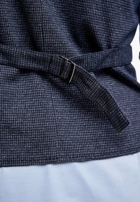 Dunkelblaues, strukturiertes Stoffkleidungsstück mit einem gewebten Muster, versehen mit einem Stoffgürtel mit einer metallischen Schnalle, übereinander getragen mit einem hellblauen Material.