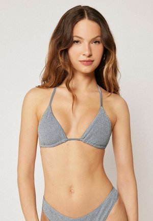 Młoda kobieta w połyskującym srebrnym bikini z długimi falującymi brązowymi włosami stoi na jasnym, jednolitym tle.